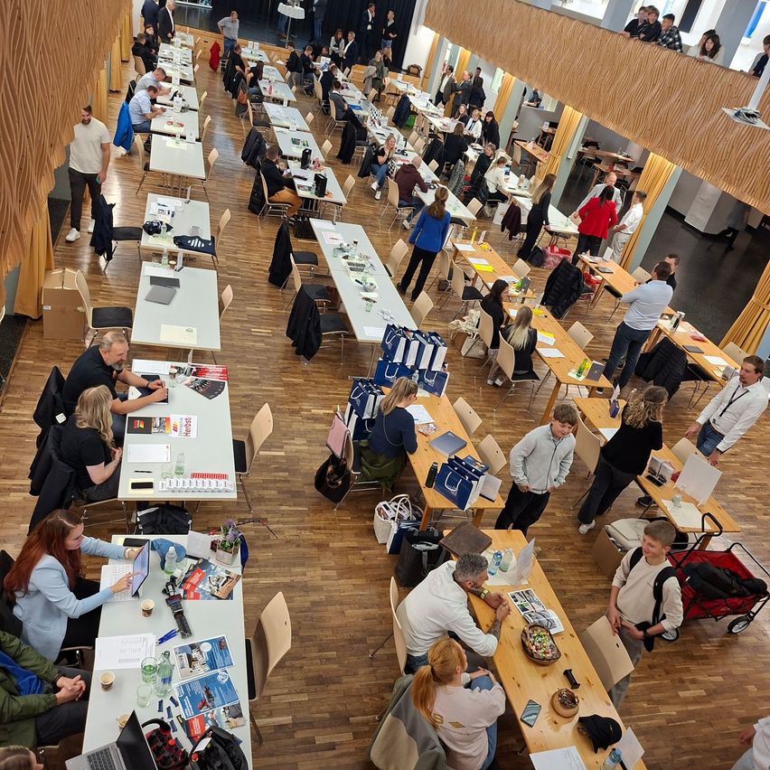 Ein großer Saal mit vielen Tischen, die jeweils von mehreren Personen besetzt sind. Einige Tische haben Laptops, Papiere und Tassen. Menschen stehen, gehen und unterhalten sich. Der Boden ist aus Holz, und die Wände sind aus Holz mit gelben Vorhängen.