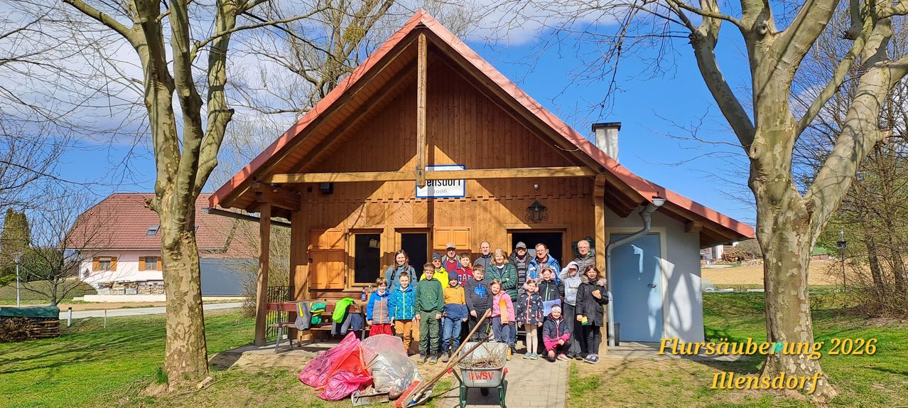 A group of people stands outside a wooden house with a red roof. Some are wearing jackets, and there are bags and a wheelbarrow nearby.