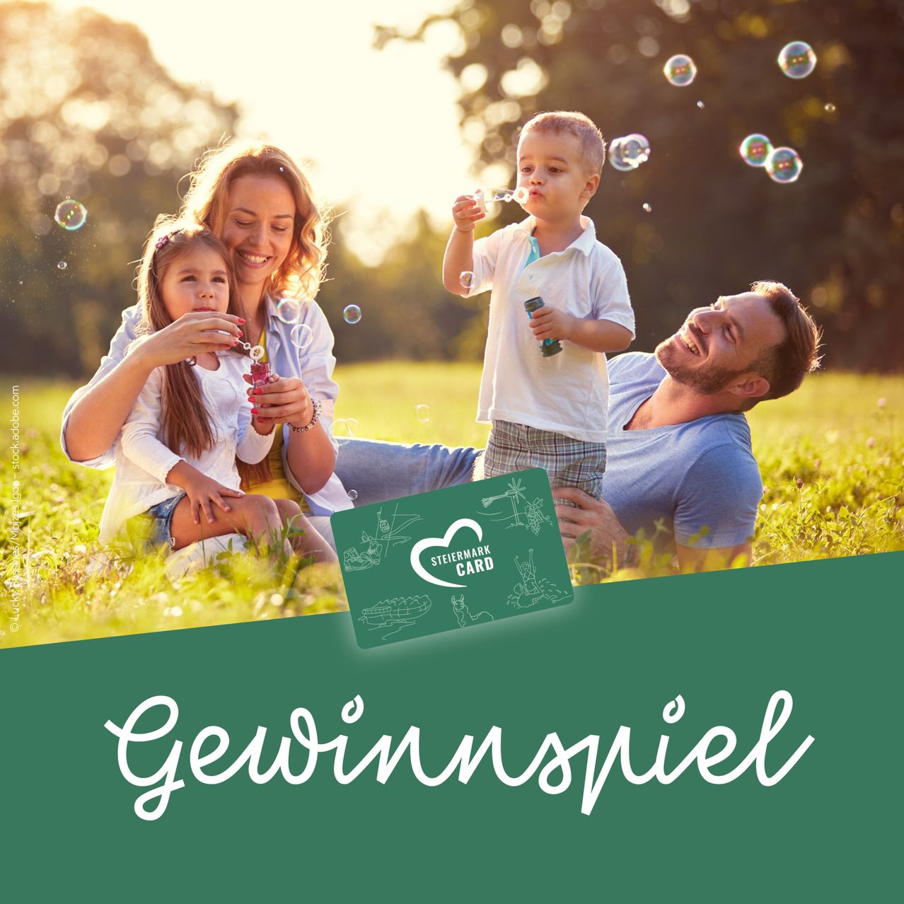 A family of four is playing in a park with bubbles. The mother and daughter are blowing bubbles while the father and son lie on the grass smiling.