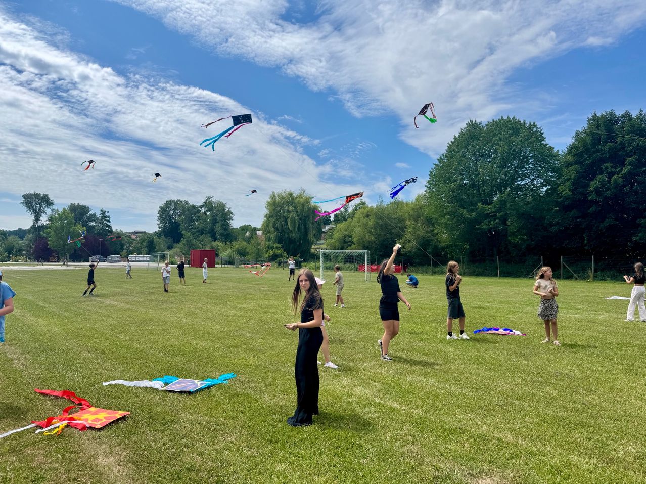 Several people fly colorful kites in a large open field under a partly cloudy sky. Some kites have landed on the ground.