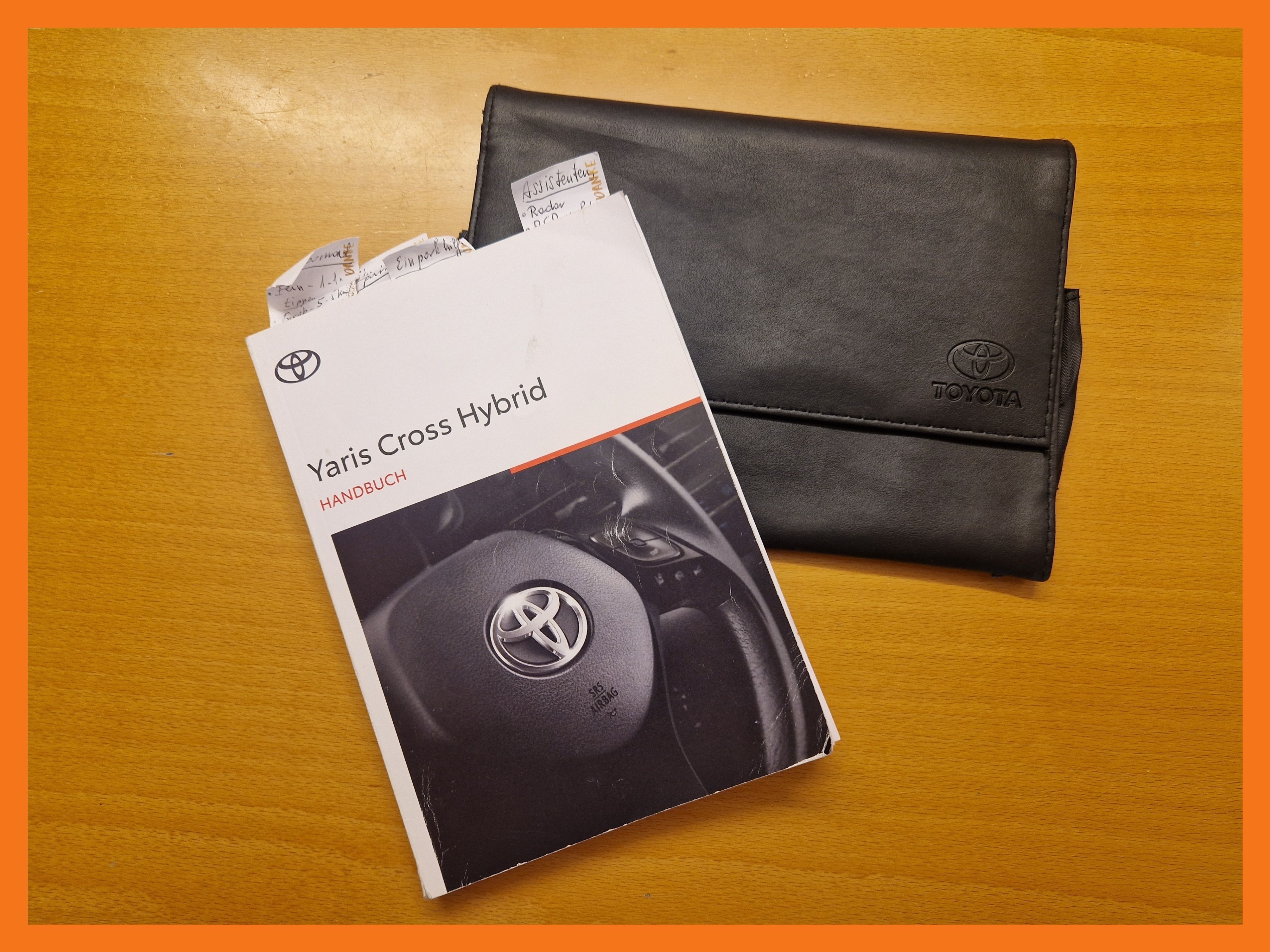 Ein Toyota Yaris Cross Hybrid Handbuch und ein schwarzer Lederetui mit Toyota Logo sind auf einer Holzoberfläche.