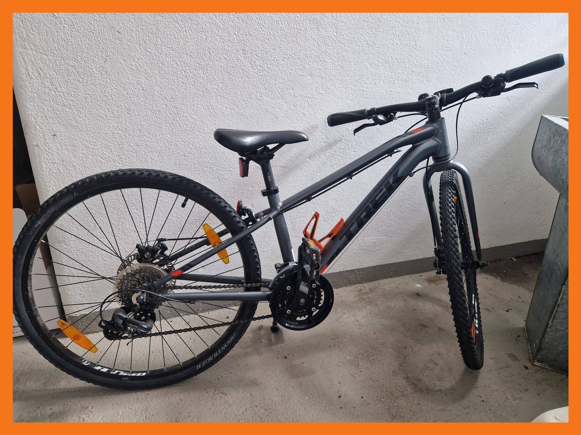 Ein graues Mountainbike mit orangen Akzenten steht gegen eine weiße Wand und zeigt seine schwarzen Lenker, Sitz und Reifen.