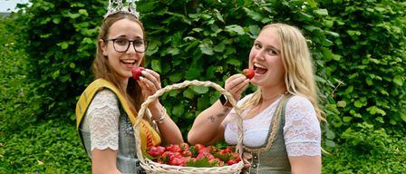 Zwei Frauen in traditioneller Kleidung halten einen Korb mit Erdbeeren. Die Frau auf der linken Seite trägt eine Krone und eine Brille. Die Frau auf der rechten Seite isst eine Erdbeere. Sie stehen vor einem Busch mit grünen Blättern.