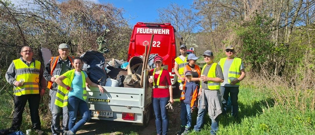 Eine Gruppe von Freiwilligen in gelben Westen steht neben einem Feuerwehrwagen, geladen mit Müll. Sie sind draußen an einem sonnigen Tag, mit Bäumen und Büschen um sie herum.