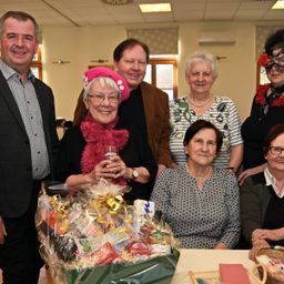 Gruppe von lächelnden Menschen, eine Frau hält einen Geschenkkorb mit einem Glas Wein