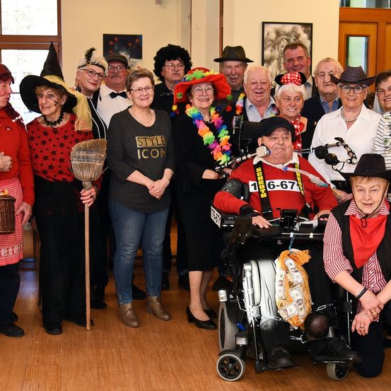 Eine Gruppe älterer Menschen ist in Kostümen gekleidet und posiert für ein Foto. Eine Frau in der Mitte hält eine Besen und trägt eine Brille. Der Mann im Rollstuhl trägt einen Hut, eine Halskette und eine Armbanduhr. Eine Frau rechts trägt einen rot-weiß karierten Schal.