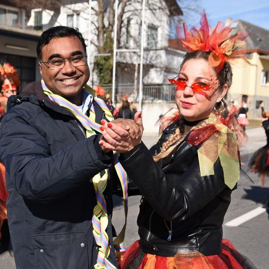 Ein Mann und eine Frau in Karnevalskostümen tanzen auf der Straße, der Mann hat bunte Bänder um den Hals und die Frau trägt eine Lederjacke und ein Tutu.