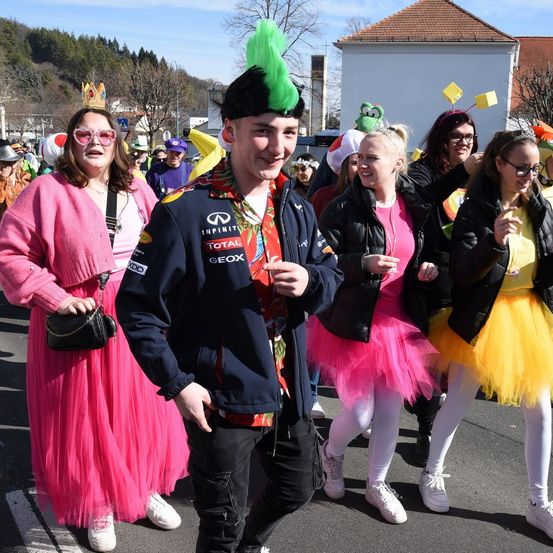 Eine Gruppe von Menschen in bunten Kostümen geht die Straße entlang. Ein Mann trägt eine blaue Jacke mit einem weißen Aufnäher und einer grünen Feder auf seinem Hut. Die Frau im rosa Kleid trägt eine Krone.