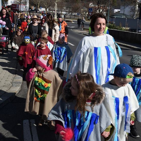 Eine Gruppe von Menschen, die auf der Straße geht, darunter eine Frau in einem weißen Gewand und mehrere Kinder in Kostümen. Einige Erwachsene tragen Hüte, und einige Leute gehen auf dem Bürgersteig. Einige Gebäude und Bäume befinden sich an den Seiten der Straße. Die Schatten der Menschen spiegeln sich auf der Straße wider.