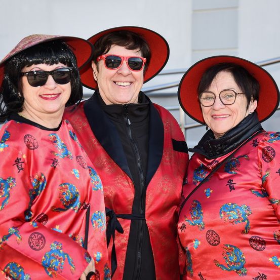 Drei Frauen mit roten Hüten und Sonnenbrillen lächeln und posieren für ein Foto vor einem weißen Gebäude.