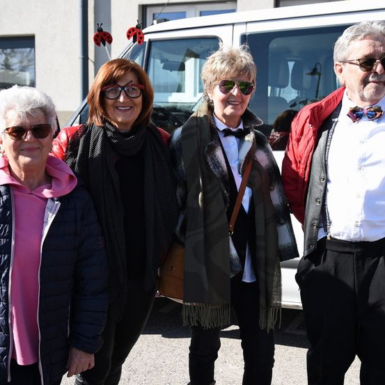 Vier ältere Erwachsene stehen neben einem weißen Van. Einer trägt eine schwarze Fliege, eine andere hat eine Krawatte. Die Frauen tragen eine Sonnenbrille und eine von ihnen ein rosafarbenes Hemd.