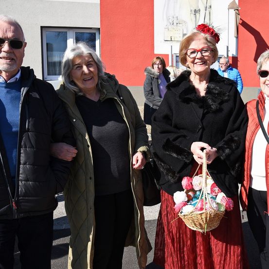 Eine Gruppe älterer Menschen, zwei Frauen und zwei Männer, stehen draußen. Eine Frau hält einen Korb mit Blumen. Die Frau in der Mitte trägt einen Pelzkragenmantel und rote Brille.