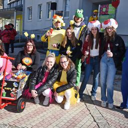 Eine Gruppe von Menschen in Kostümen posiert für ein Foto. Sie sind draußen in der Nähe eines Gebäudes. Einige Leute tragen Brillen und Hüte. Eine Frau vorne hält einen roten Wagen mit Spielzeug.