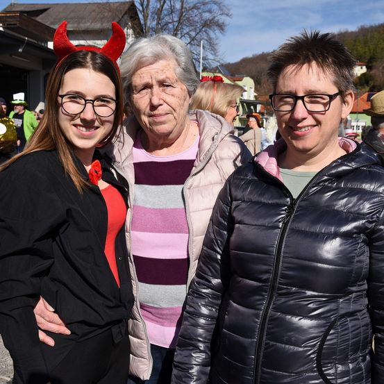Drei Frauen stehen eng zusammen vor einem verschwommenen Hintergrund aus Gebäuden und Bergen. Die Frau auf der linken Seite trägt ein Teufelshörner-Kopfband und einen schwarzen Mantel. Die Frau in der Mitte hat graues Haar und trägt ein rosa-grau gestreiftes Hemd. Die Frau auf der rechten Seite trägt eine schwarze, gefütterte Jacke.