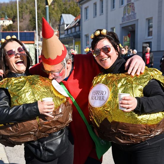Drei Personen sind in goldener Folienkleidung gekleidet, zwei als Schokoladenei und einer als Bleistift, und lächeln für ein Foto bei einer Veranstaltung im Freien.