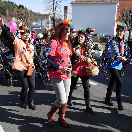 Fünf Frauen in bunten Kostümen laufen während eines Umzugs auf der Straße, einige halten Körbe und andere tragen einen rosa Luftballon.