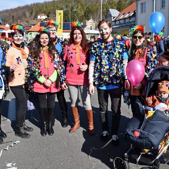 Eine Gruppe von Menschen in bunten Kostümen und mit Gesichtsbemalung steht auf einer Straße mit Luftballons. Ein Baby ist in einem Kinderwagen rechts.