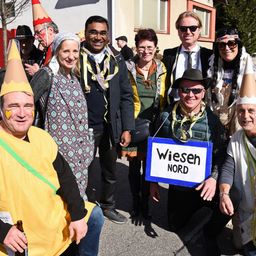 Eine Gruppe von Menschen in Kostümen posiert für ein Foto vor einem Gebäude. Ein Mann in einem gelben Shirt hält ein Bier, während andere Hüte und Schals tragen.