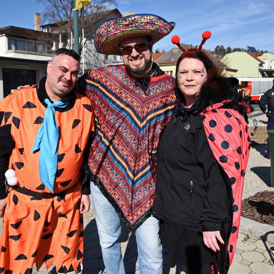Drei Menschen in Kostümen stehen nebeneinander und lächeln für die Kamera. Der Mann auf der linken Seite ist als Fred Flintstone verkleidet und die Frau auf der rechten Seite als Marienkäfer.