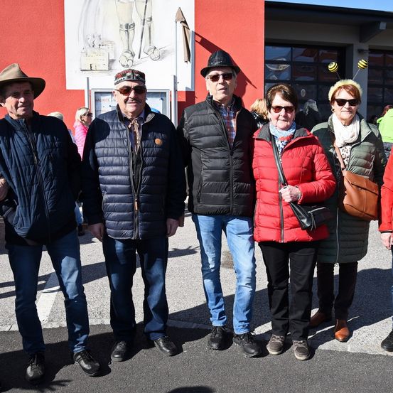 Fünf Personen in Jacken und mit Sonnenbrillen stehen vor einem roten Gebäude. Der Mann auf der linken Seite trägt einen Hut.
