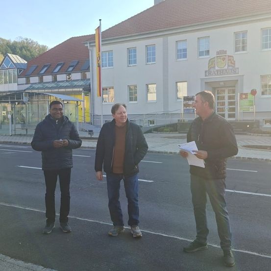 Drei Männer stehen auf der Straße vor einem Gebäude mit rotem Dach. Ein Mann hält ein Papier.