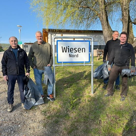 Vier Männer stehen neben einem Schild für Wiesen Nord. Sie halten Müllsäcke.