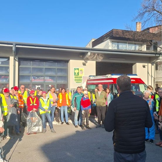 Gruppe von Menschen in Sicherheitswesten und Mützen steht an einem sonnigen Tag vor einer Feuerwache.