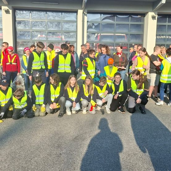 Eine Gruppe von Kindern in gelben Warnwesten kniet vor einem Gebäude. Sie halten ein Werkzeug und scheinen an einer Reinigungsaktion teilzunehmen.