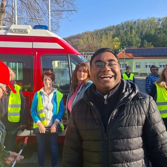 Ein Mann mit einem Lächeln und Brille steht vor einem roten Krankenwagen. Hinter ihm steht eine Frau in einer gelben Weste mit einer weiteren Frau in einem rosa Oberteil. Im Hintergrund sind mehrere Personen in Sicherheitswesten zu sehen.