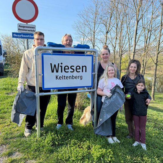 Eine fünfköpfige Familie steht hinter einem Wiesen Keltenberg-Schild, hält Taschen und lächelt für ein Foto. Sie tragen Handschuhe und stehen auf Gras mit Bäumen im Hintergrund.