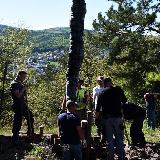 Eine Gruppe von Männern arbeitet im Freien an einem Projekt. Ein Mann benutzt eine Schaufel, während andere eine Spitzhacke verwenden. Sie befinden sich in einem Wald mit Bäumen und einem Holzzaun.