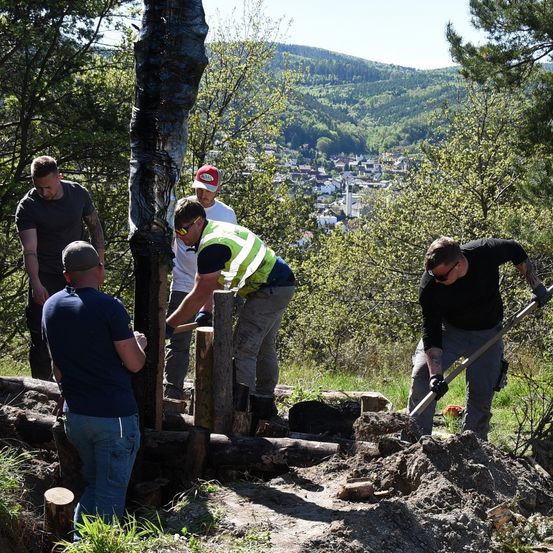 Fünf Männer im Wald arbeiten gemeinsam daran, mit einem Spaten zu graben und auf Baumstämme zu schlagen. Sie sind von Bäumen und grünen Pflanzen umgeben, und in der Ferne gibt es eine Stadt mit Gebäuden und einem Berg.