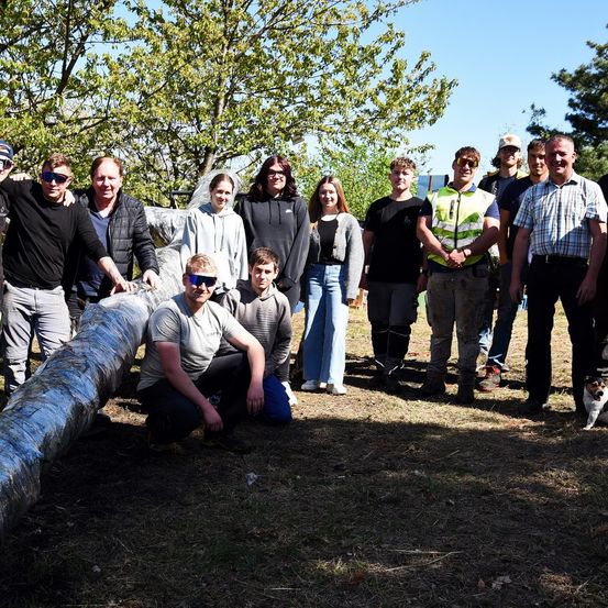 Eine Gruppe von Menschen steht und kniet in einem Park mit einem großen Objekt vor ihnen. Die Person vorn trägt eine graue Jacke und schwarze Hose.