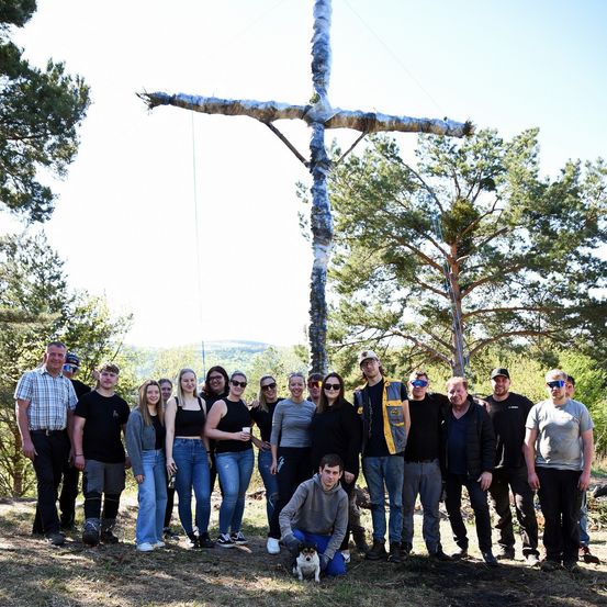 Eine Gruppe von Menschen in Sonnenbrillen und Turnschuhen steht vor einem großen Holzkreuz. Das Kreuz ist in Silberfolie gewickelt und steht auf einem Rasen.