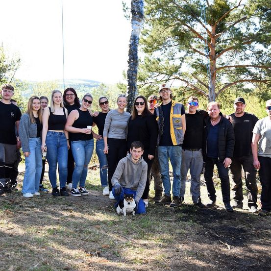 Eine Gruppe von Menschen, die in einem Wald für ein Foto posiert und einen kleinen Hund vor sich stehen.