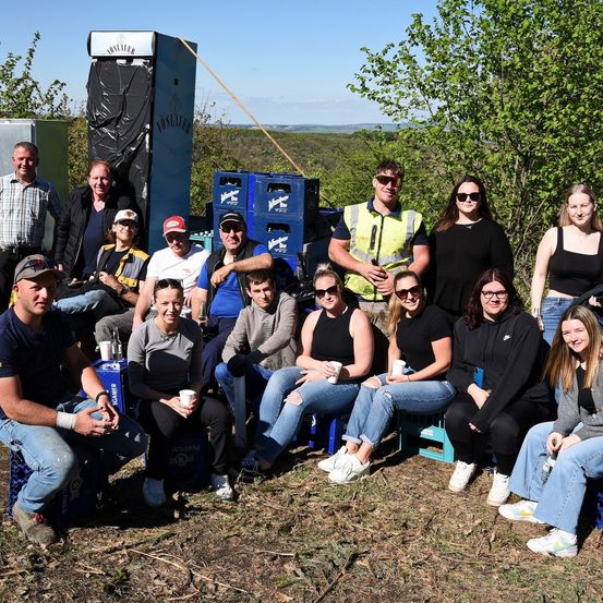 Eine Gruppe von Menschen posiert für ein Foto in einem Außenbereich. Dahinter befinden sich blaue Kisten und ein Holzpfahl. Die Menschen tragen Freizeitkleidung, und einige tragen eine Brille. Einer von ihnen trägt eine Sicherheitsweste. Sie stehen und sitzen auf blauen Stühlen, und einige halten Tassen. In der Ferne gibt es Bäume und einen Berg.