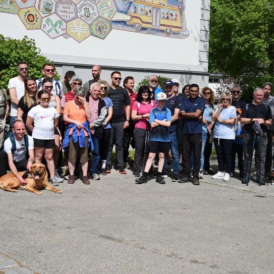 Eine Gruppe von Menschen mit einem Hund vor einem Gebäude mit einer Mosaikwand.