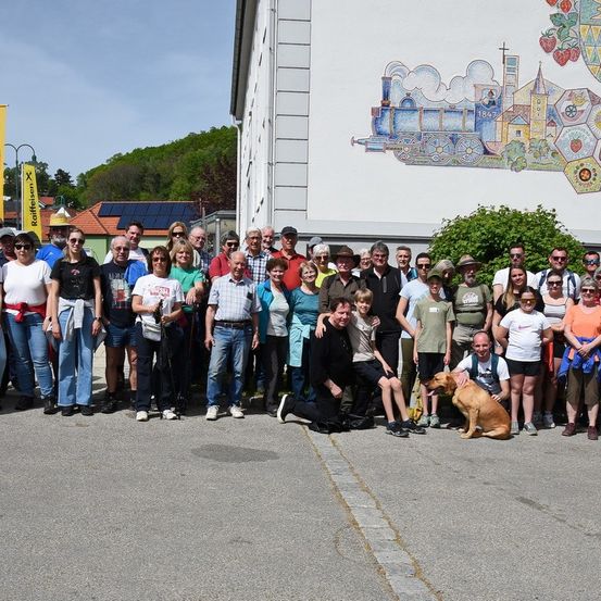 Eine große Gruppe von Menschen in Freizeitkleidung und mit Sonnenbrillen, einige mit Hüten, posiert für ein Foto vor einem Gebäude mit einem Wandbild.
