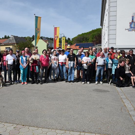 Eine Gruppe von Menschen in verschiedenen farbigen Shirts und Accessoires posiert für ein Foto vor einem Gebäude mit Fahnen und Solarpanelen im Hintergrund.