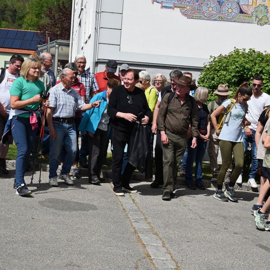 Eine Gruppe von Menschen steht vor einem Gebäude mit einem Wandbild. Sie tragen Freizeitkleidung, einige haben Sonnenbrillen und eine Frau hält einen Gehstock.