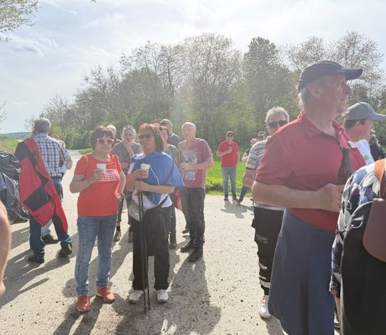 Eine Gruppe von Menschen, einige mit Hüten und Brillen, stehen auf einem Feldweg mit Bäumen im Hintergrund.
