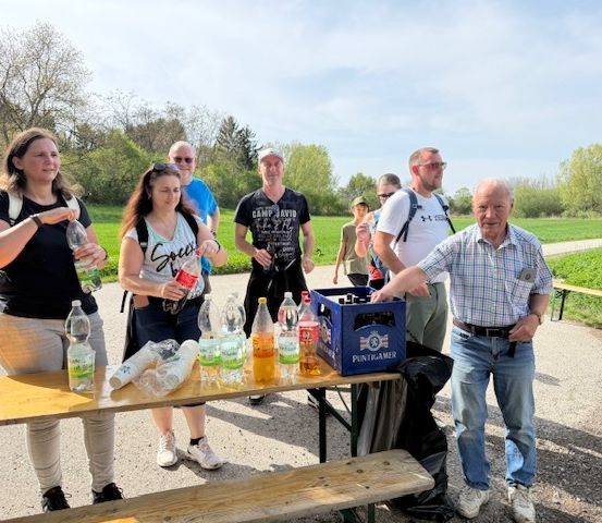 Mehrere Personen stehen um einen Picknicktisch. Eine Frau in Schwarz hält eine Flasche und ein Mann lächelt.
