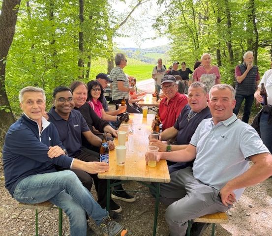 Eine Gruppe von Menschen sitzt um einen langen Holztisch in einem Wald. Sie haben Getränke und Essen auf dem Tisch.