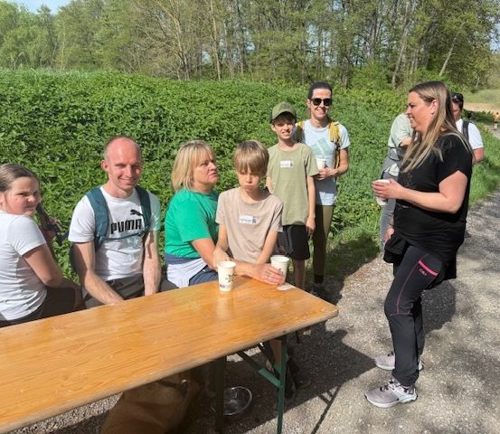 Eine Gruppe von Menschen, einschließlich Erwachsener und Kindern, sitzt um einen Holztisch mit Tassen herum im Freien.