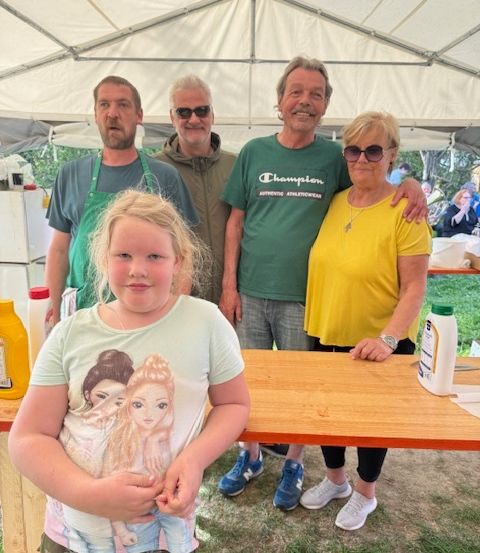 Fünf Personen stehen in einem Zelt mit einem Tisch und Milchflaschen. Der Mann in der Mitte trägt ein grünes Shirt.