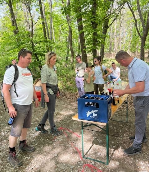 Eine Gruppe von Menschen versammelt sich in einer Waldlichtung. Sie stehen um einen Tisch, auf dem Flaschen und Dosen stehen, wahrscheinlich bei einem Picknick.