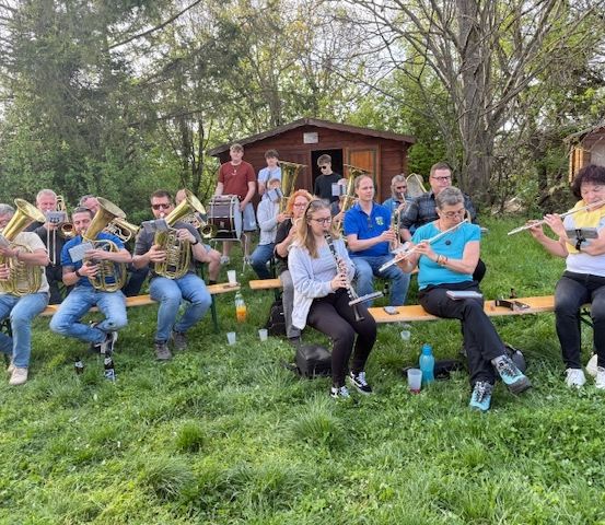 Eine Gruppe von Musikern sitzt auf Bänken in einem Grasbereich, einige halten Musikinstrumente. Dahinter ist ein kleines Holzhaus und Bäume zu sehen.