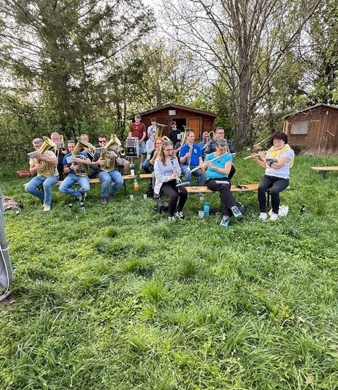 Eine Gruppe von Musikern sitzt auf einer Bank im Gras und hält ihre Musikinstrumente. Sie spielen wahrscheinlich in einem Park.