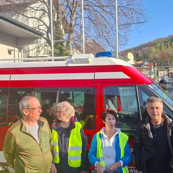 Vier Menschen stehen vor einem roten Feuerwehrauto. Sie tragen Warnwesten. Die Frau in der Mitte hat den Mund offen. Das Gebäude hinter ihnen hat einen Zaun und einen Baum.
