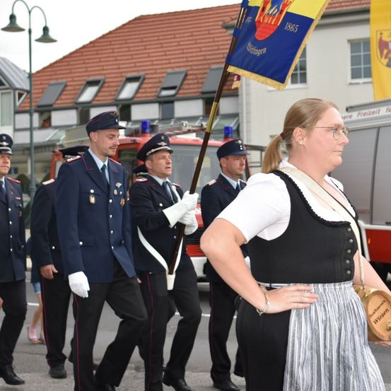 Eine Gruppe von Feuerwehrleuten marschiert mit einer Fahne vor einem Gebäude mit rotem Dach.
